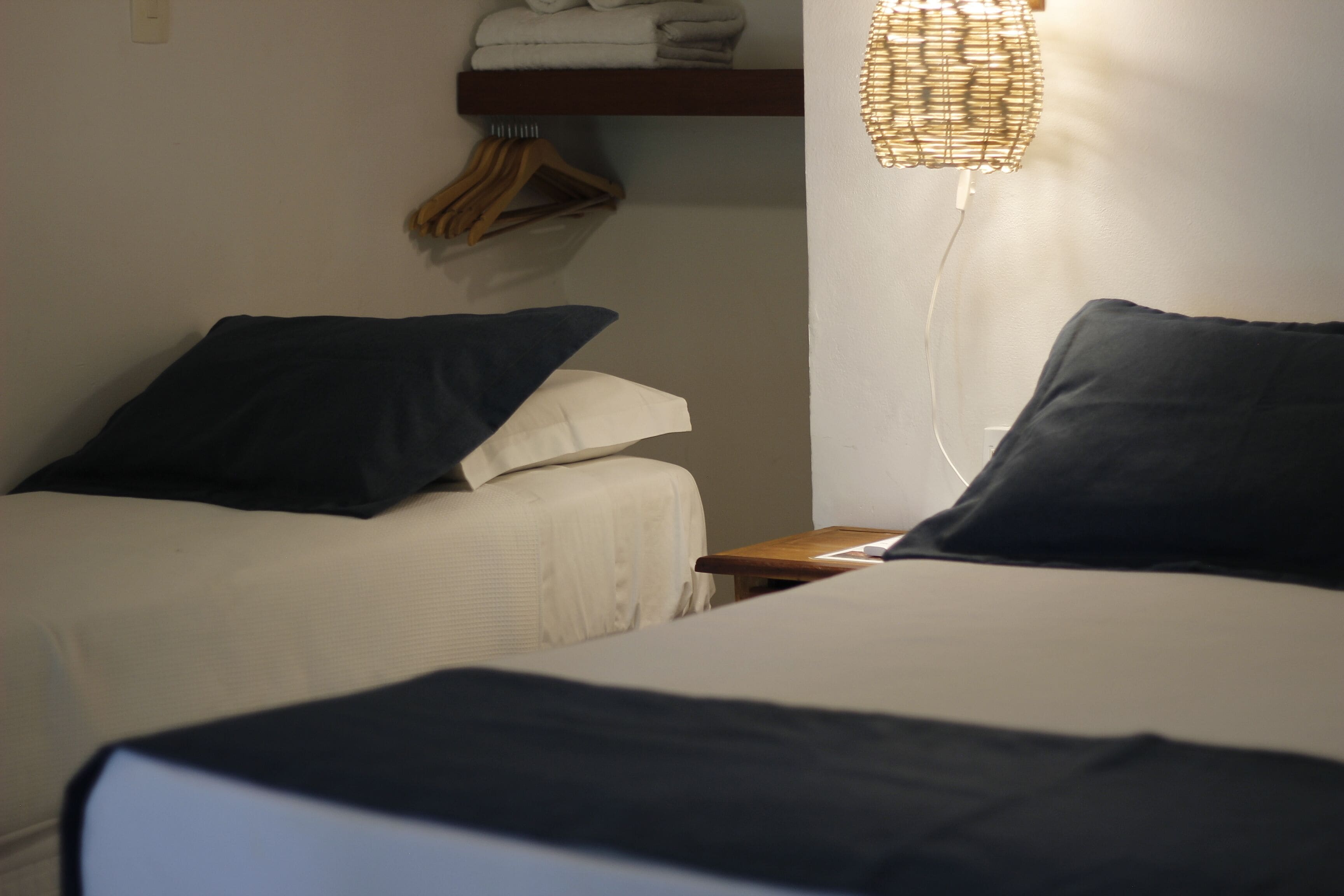 A hotel room with two beds, a bedside table, a hanging clothes rack, and a wall-mounted shelf with folded towels.