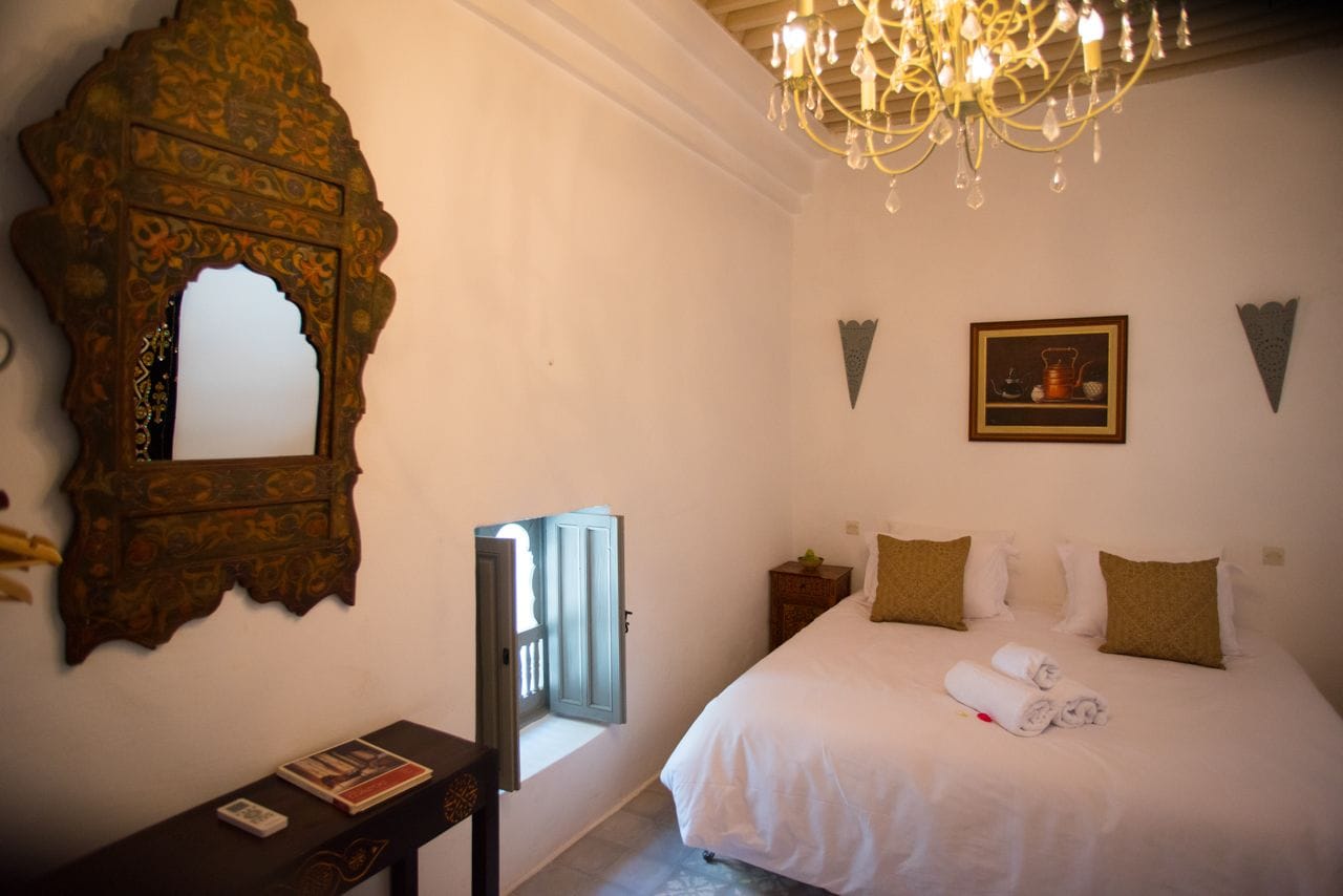 A bedroom with a large bed, a wooden mirror, a chandelier, and a framed picture on the wall.