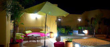 Un patio exterior con una cama, una mesa y sillas, rodeado de plantas en macetas y un paraguas grande.