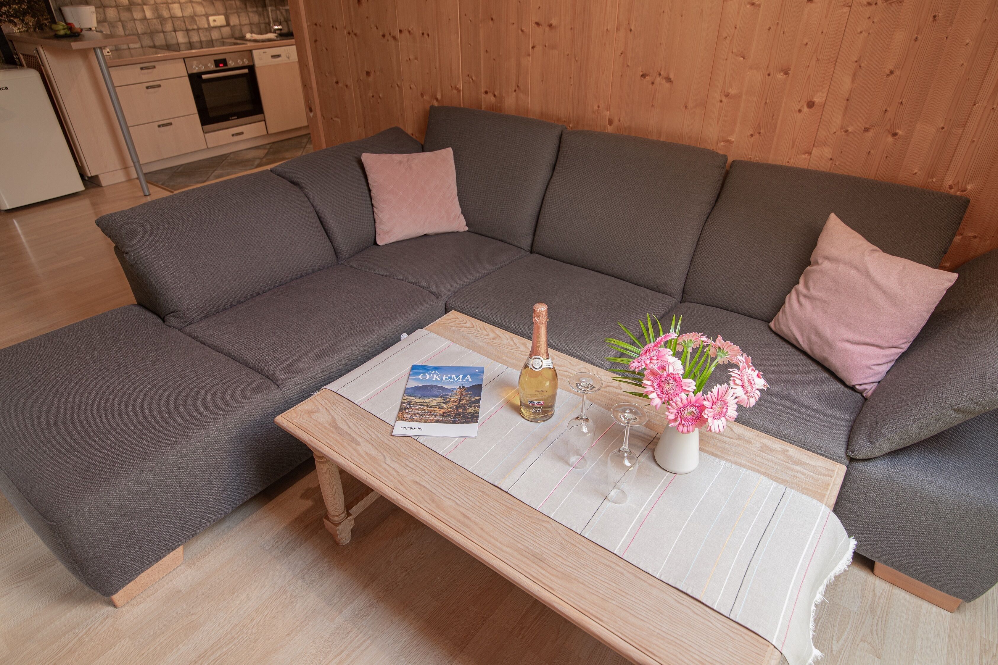 A modern living room with a grey sectional sofa, a wooden coffee table with a magazine, a bottle of wine, and a vase of flowers.