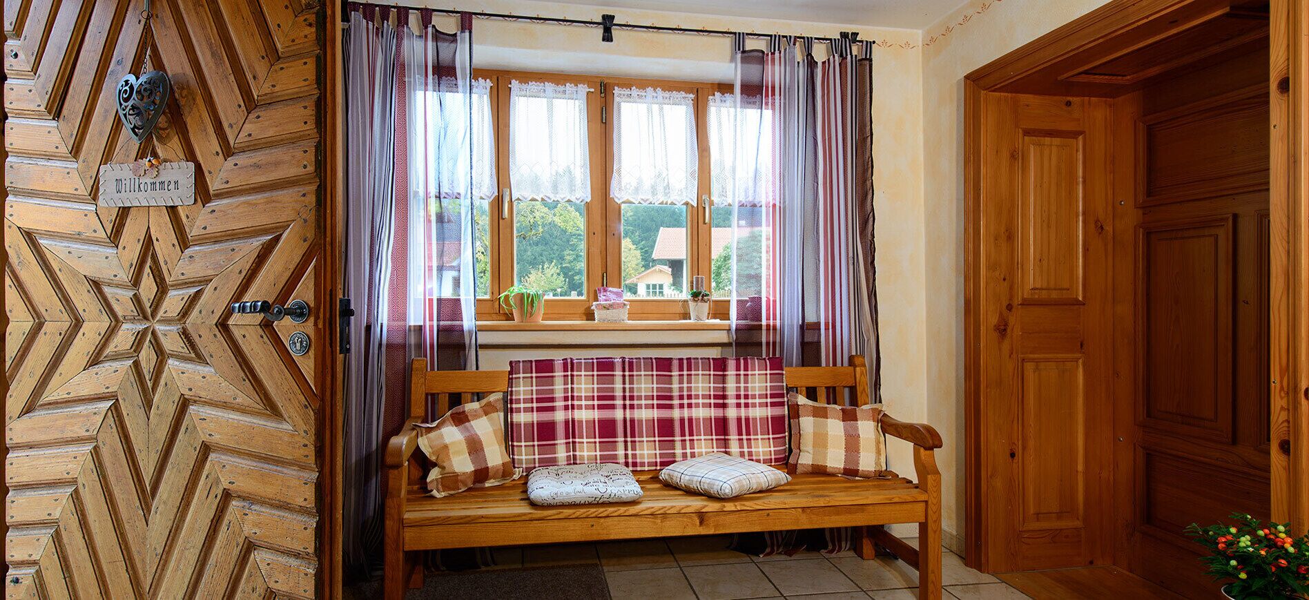A wooden bench with plaid cushions, a window with lace curtains, and a 'Willkommen' sign on the wall.