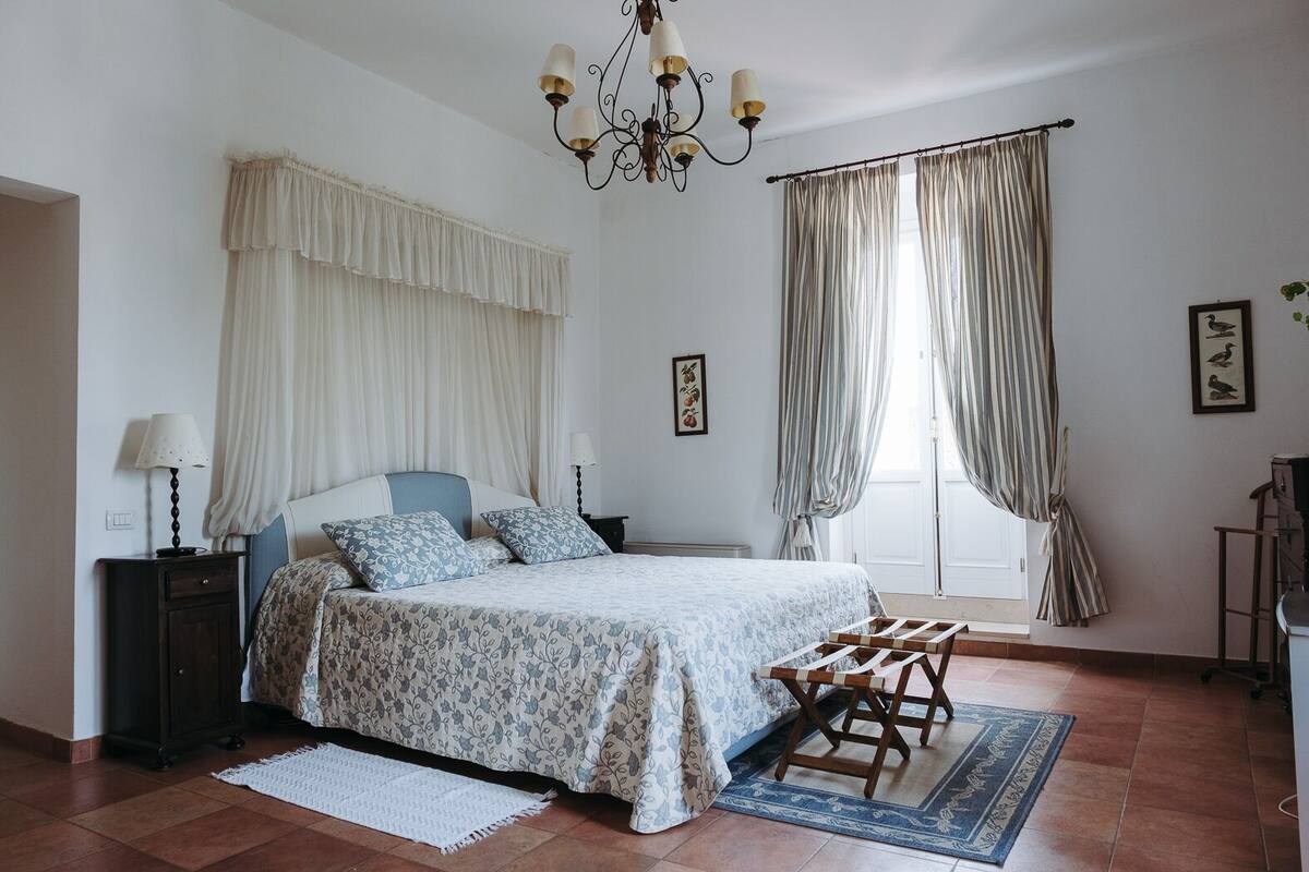 A bedroom with a bed, bedside tables, a chandelier, curtains, and a view of the outdoors.