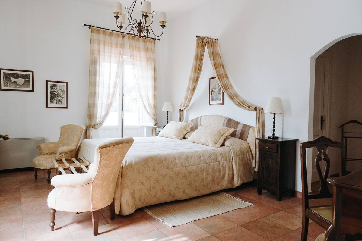 A bedroom with a bed, two bedside tables, a chair, a radiator, and a window with curtains.