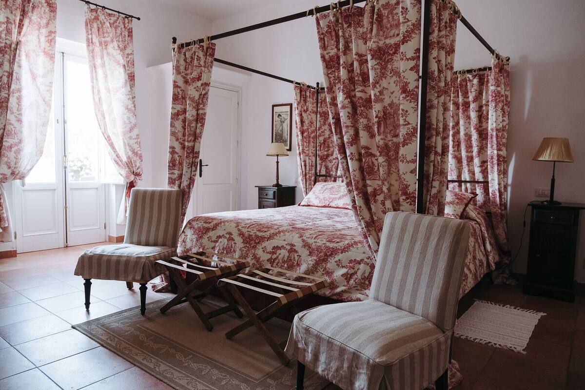 A four-poster bed with floral curtains, a wooden chair, a bedside table with a lamp, and a rug on the floor.