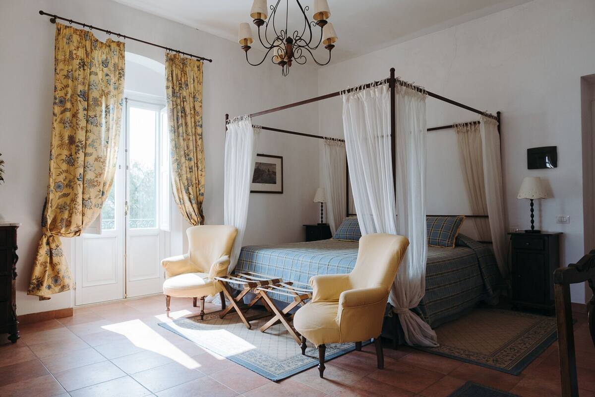A four-poster canopy bed with a blue and white checkered bedspread, two armchairs, a nightstand with a lamp, and a chandelier.