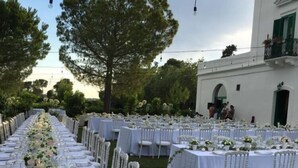 A long outdoor dining area with white tables and chairs, surrounded by greenery and a building in the background. - Golf Club Metaponto (Bernalda)