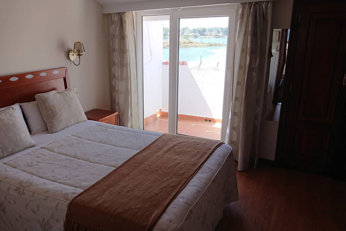 A hotel room with a bed, a nightstand, a wall-mounted lamp, and a view of a pool through a sliding glass door.
