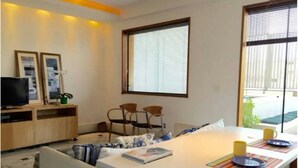 A modern living room with a large white table, a TV on a wooden stand, and a window with blinds. - Copa Loft (Rio de Janeiro)