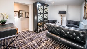 A modern living room with a black leather sofa, a patterned carpet, a black cabinet with glass doors, and framed artwork on the wall. - The Royal Standard (Ely)