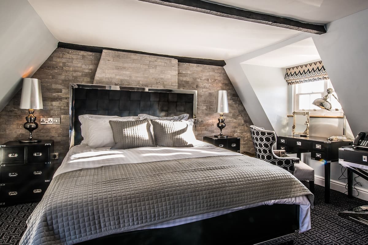 Un dormitorio moderno con una cama grande, un cabecero oscuro y un techo inclinado. Hay dos mesitas de noche con lámparas y un escritorio con una silla.