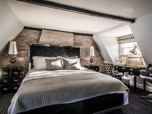 A modern bedroom with a large bed, a dark headboard, and a sloped ceiling. There are two bedside tables with lamps and a desk with a chair. - The Royal Standard (Ely)