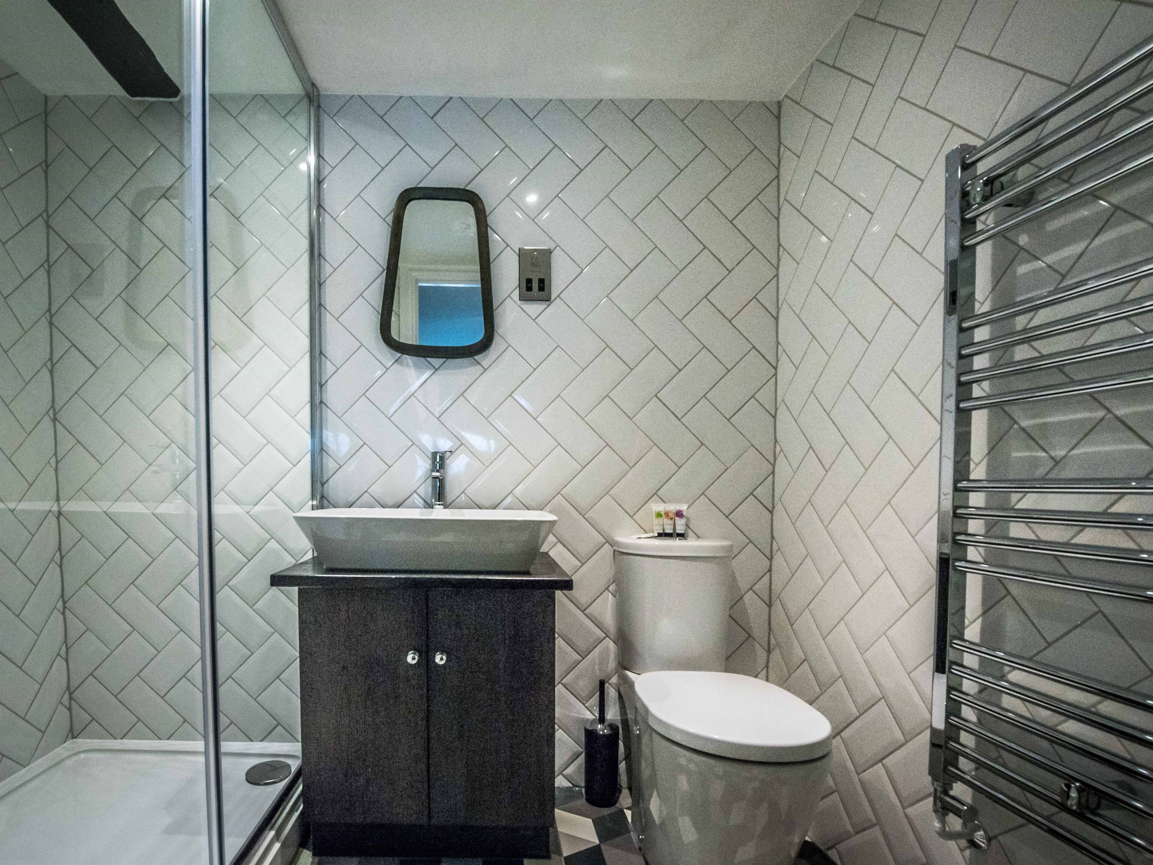 A modern bathroom with a herringbone tile pattern, a glass shower enclosure, a dark cabinet, a white toilet, and a towel rack.