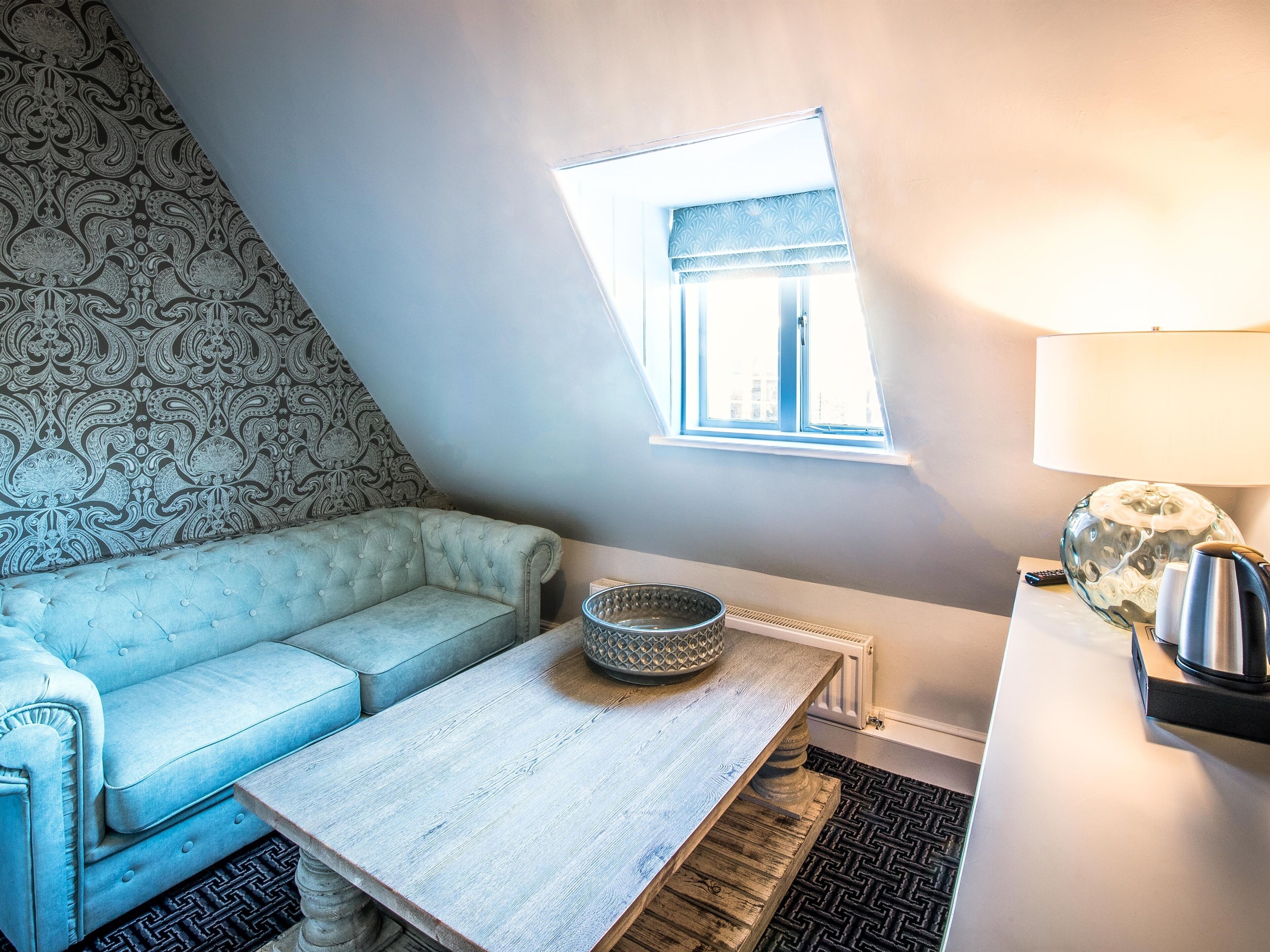 A cozy room with a light blue sofa, a wooden table, a patterned wall, a small window, and a lamp.