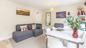 A living room with a grey sofa, a dining table with white chairs, and a red vase with flowers. - Greyhound Road Rooms (London)