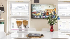 A modern dining area with a white table, chairs, a vase with flowers, a remote control, and a menu stand. - Greyhound Road Rooms (London)