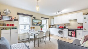 A modern kitchen with white cabinets, a built-in oven, and a washing machine. There is a dining table with chairs and a window with blinds. - Greyhound Road Rooms (London)
