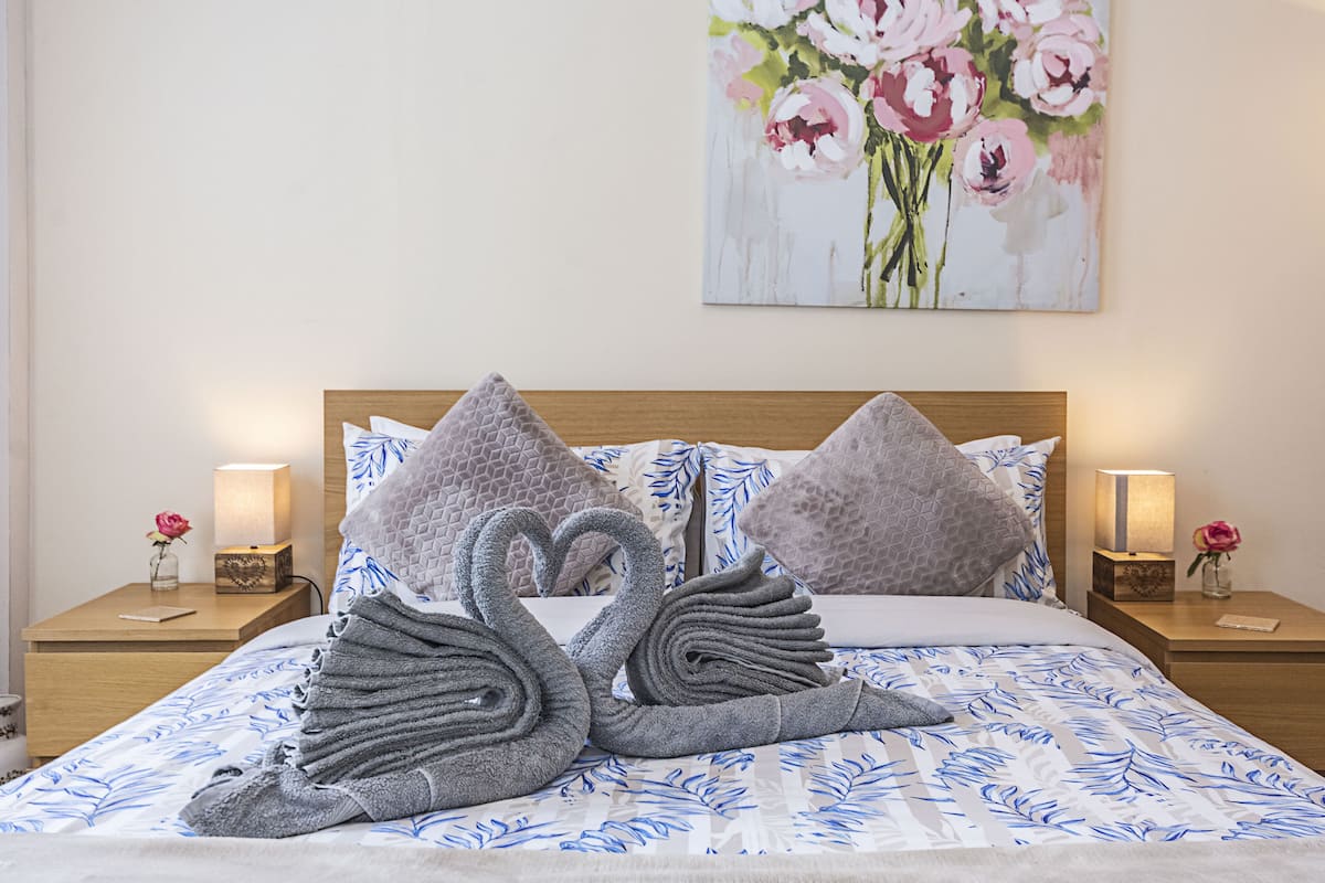 Ein ordentlich bezogenes Bett mit einem blumigen Bettüberwurf, zwei Kissen und zwei schwanenförmigen Handtüchern. Auf jedem Nachttisch stehen eine Nachttischlampe und eine Vase mit einer Blume.