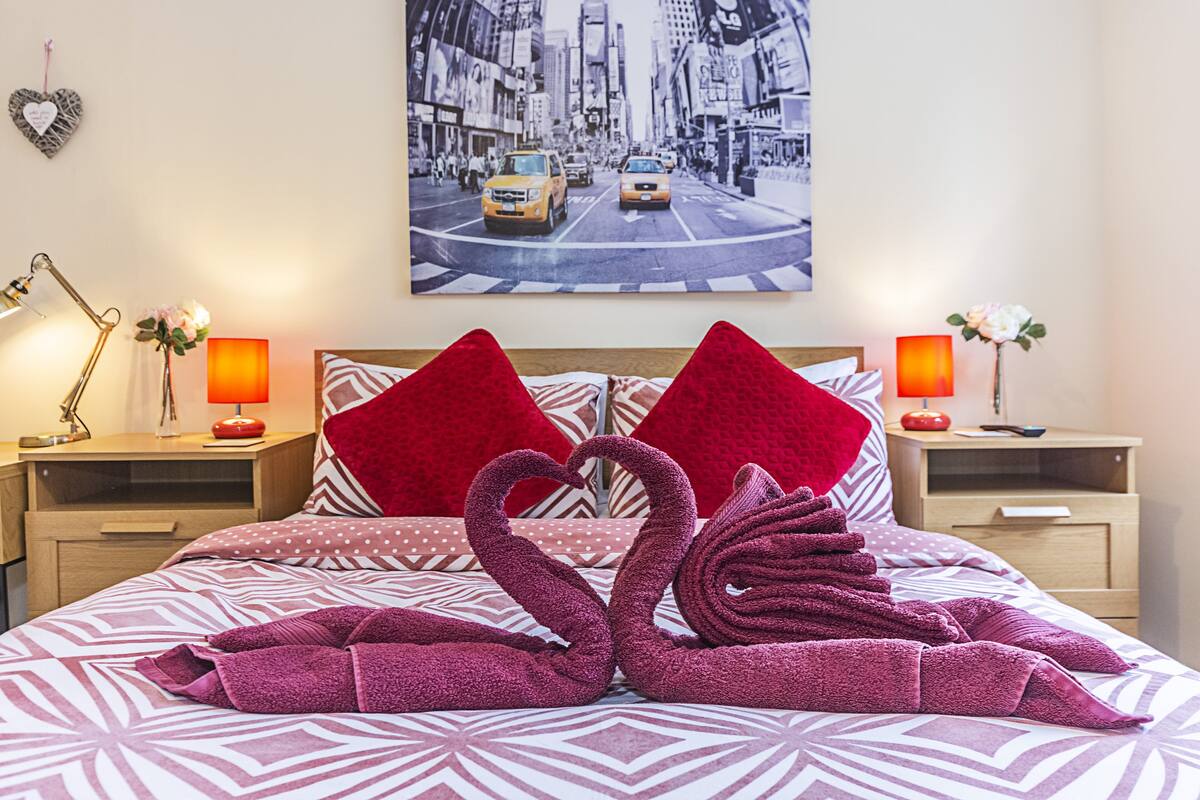 Ein Hotelzimmer mit einem Bett, das eine geometrische Bettdecke, zwei rote Kissen und eine herzförmige Handtuchanordnung zeigt. An der Wand hängt ein gerahmtes Stadtbild, und es gibt zwei Nachttische mit Lampen und kleinen Vasen.