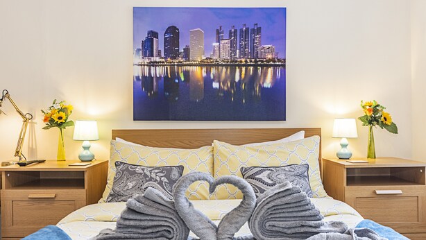 A hotel room with a bed, two nightstands, a cityscape painting, and towels arranged in swan shapes. - Greyhound Road Rooms (London)