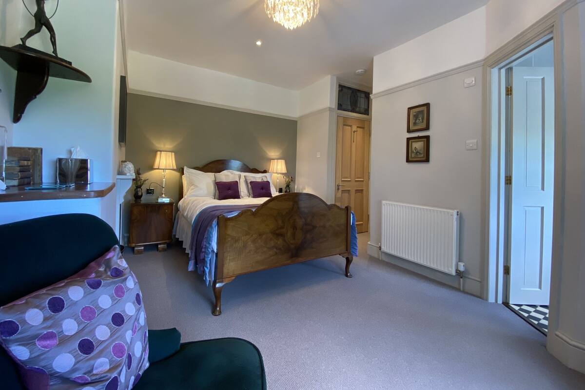 A bedroom with a wooden bed, two bedside lamps, a chandelier, a radiator, and a door leading to another room.