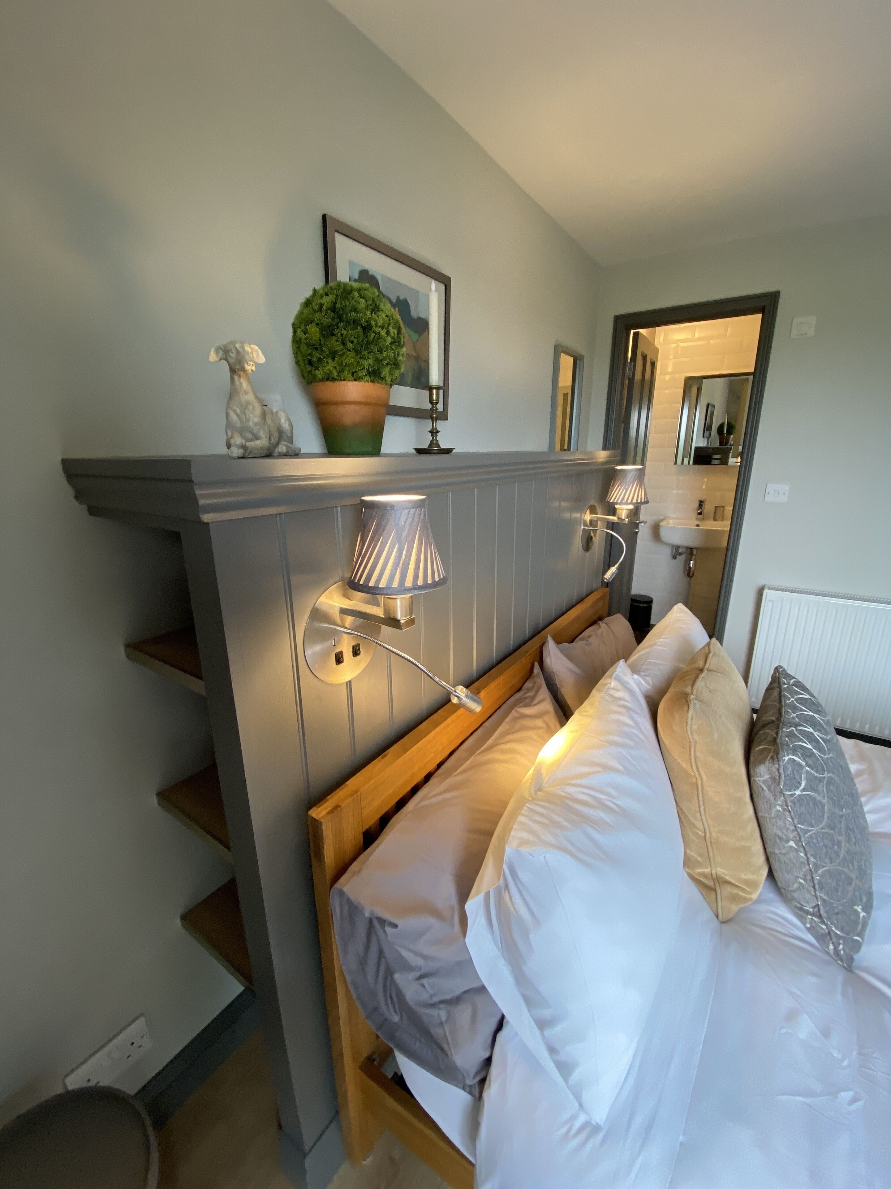 A bedroom with a bed, a nightstand, a lamp, a mirror, a potted plant, and a framed picture on the wall.