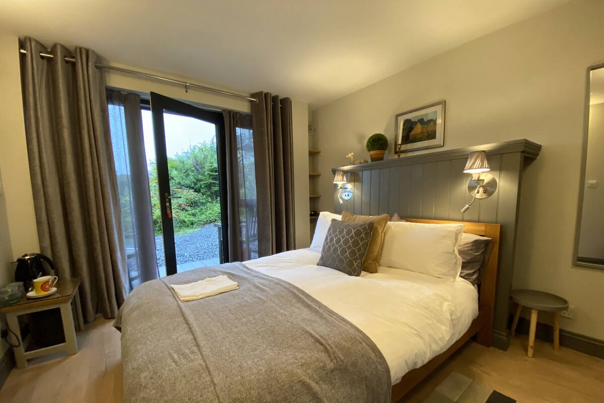 A hotel room with a large bed, a bedside table with a lamp, a view of greenery outside, and a painting on the wall.