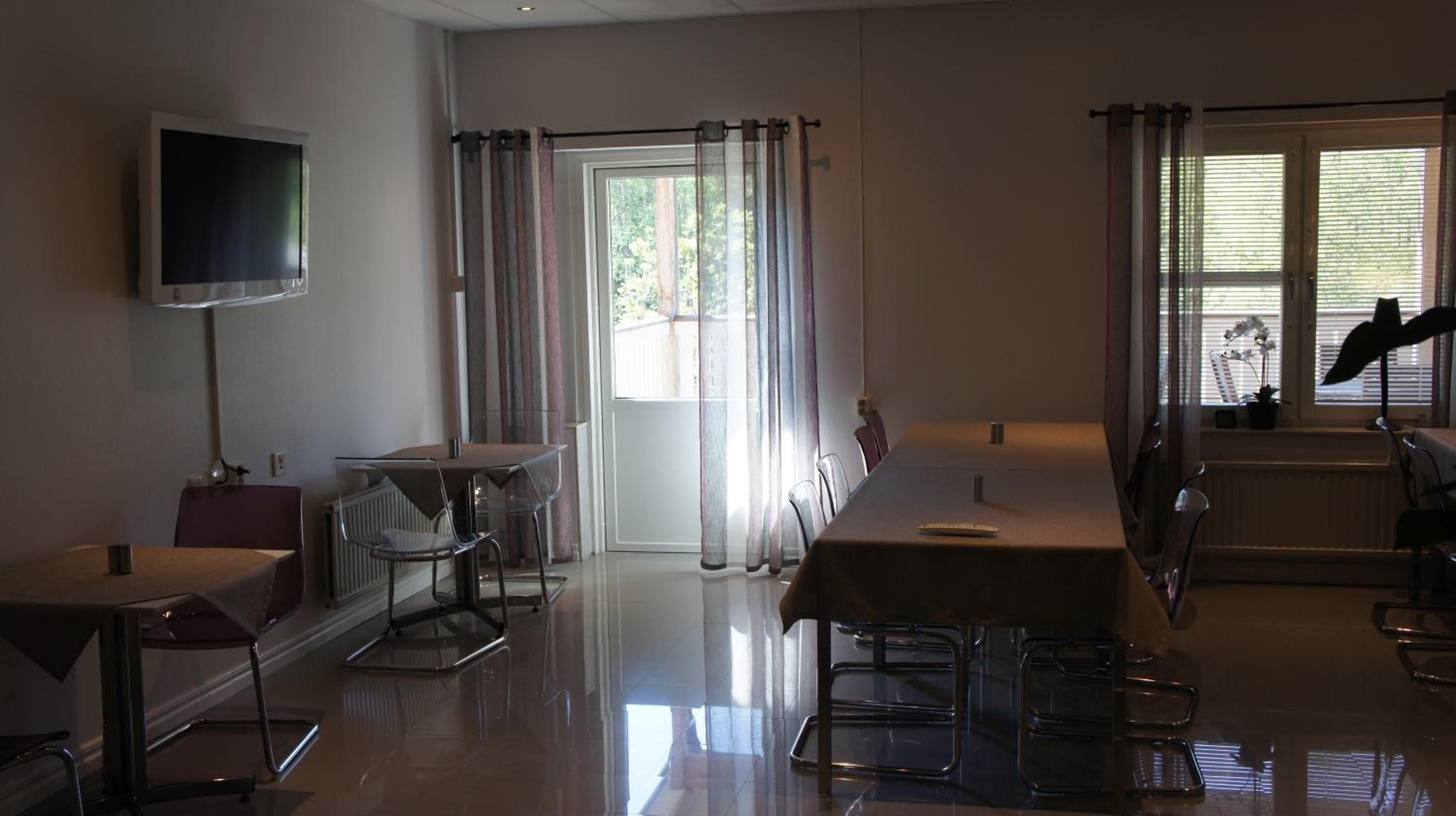 Una habitación amplia con una gran televisión de pantalla plana montada en la pared, una mesa de comedor con sillas, un sofá y una ventana con cortinas.