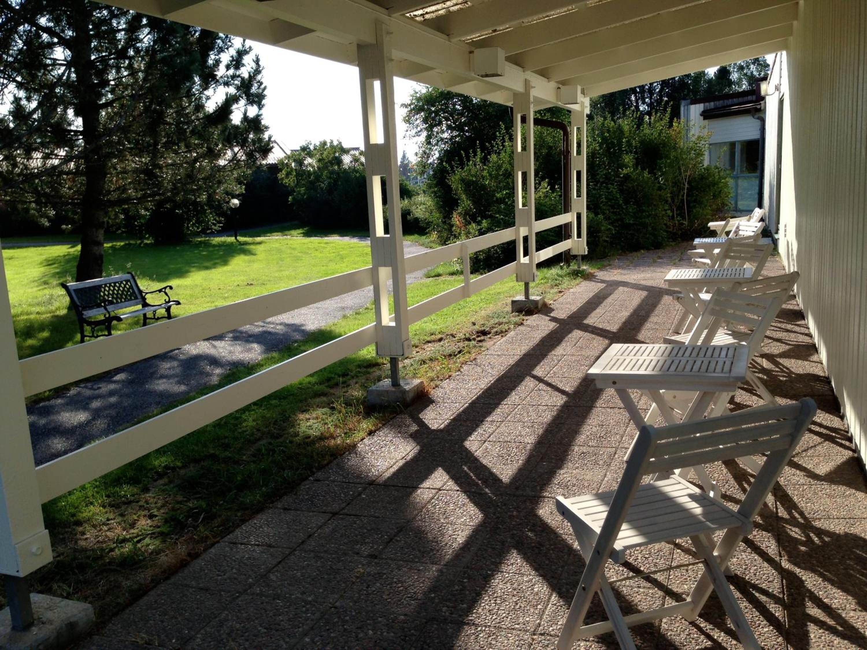 A covered outdoor seating area with white chairs and a bench, overlooking a grassy park with trees.