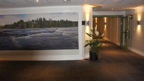 A hallway with a large wall mural of a river landscape, a potted plant, and a door with a sign indicating room rates. - Hotell Vindelngallerian (Vindeln)