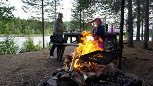 A campsite with a fire pit, a table with food, and a river in the background. - Hotell Vindelngallerian (Vindeln)