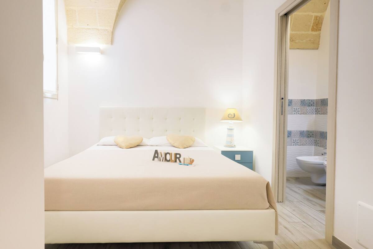 Una cama con cabecera, una lámpara de noche y un cojín decorativo con la palabra 'AMOUR'.