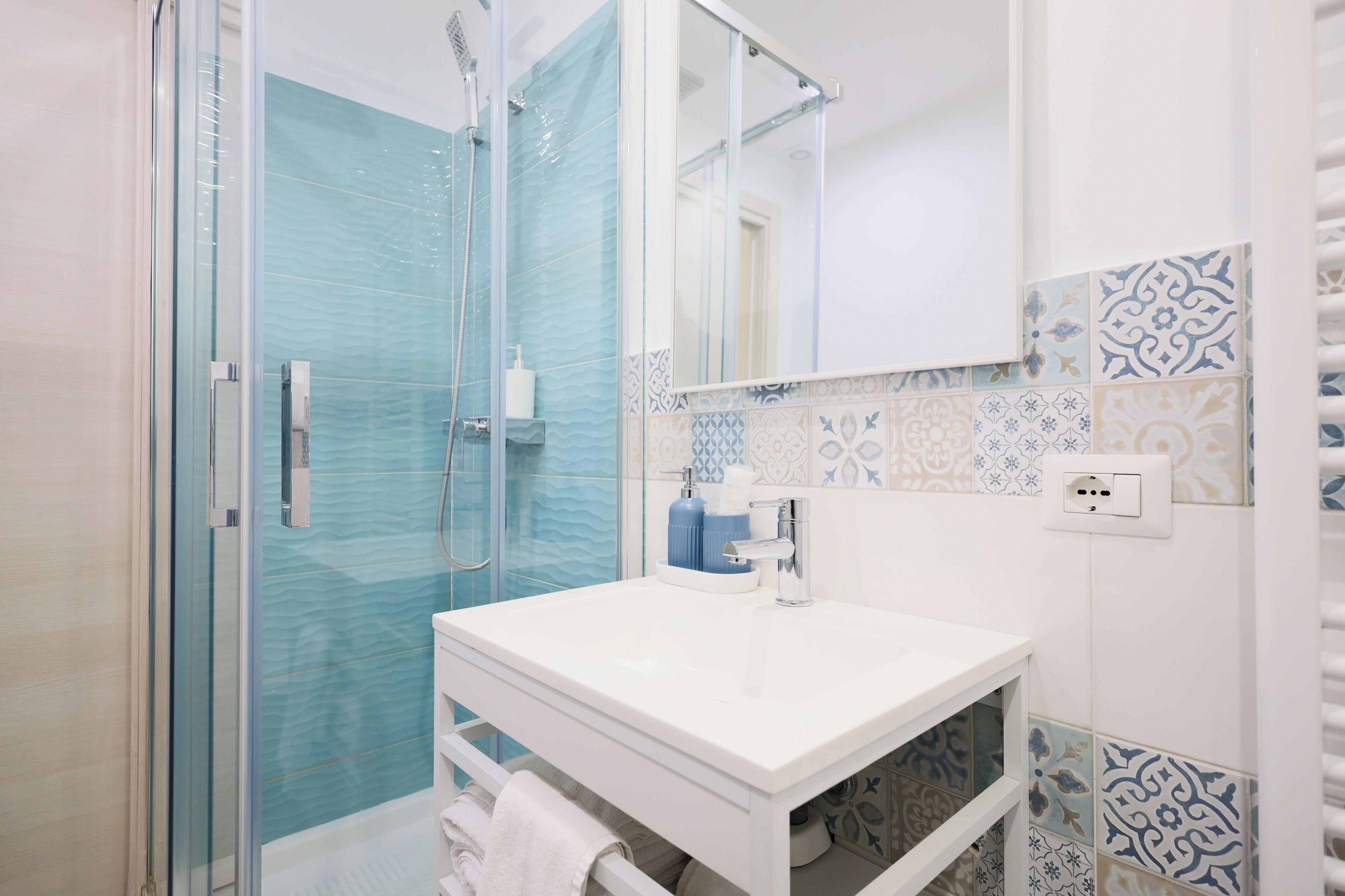 A bathroom with a glass shower enclosure, a white sink with a mirror, and patterned tiles on the walls.