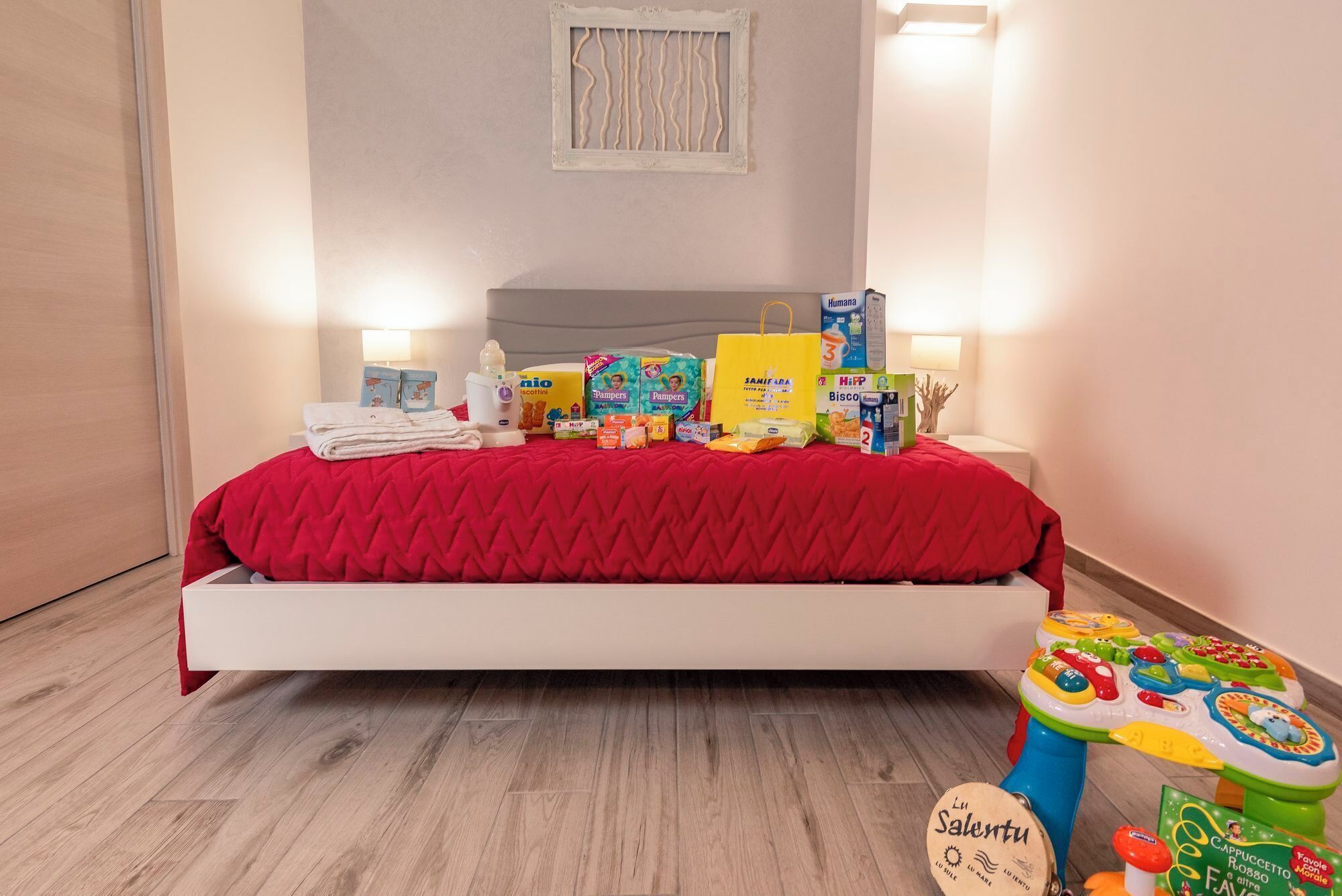 Ein Schlafzimmer mit roter Bettdecke, weißem Kopfteil und Holzboden. Auf dem Bett liegen verschiedene Gegenstände, darunter eine gelbe Tasche, eine Windelbox und eine Babyflasche. Neben dem Bett ist ein Spielzeug zu sehen.