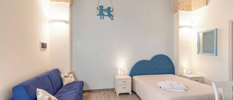 A bedroom with a blue sofa, a bed with a blue headboard, a nightstand with a lamp, and a wall-mounted decoration.