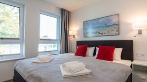 Ein ordentlich bezogenes Bett mit weißen und roten Kissen, ein Strandbild an der Wand und ein Fenster mit Vorhängen.