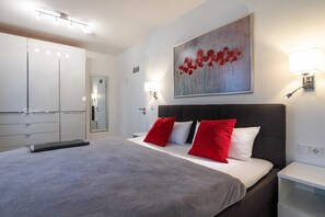 A modern hotel room with a large bed, a grey blanket, red and white pillows, a painting of red flowers, a bedside lamp, and a wardrobe.