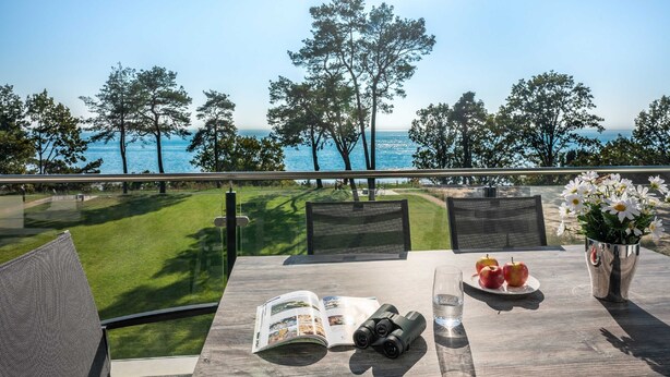 Ein Tisch mit einer Zeitschrift, Ferngläsern, Äpfeln und einem Glas Wasser, mit Blick auf eine Wiese und das Meer.