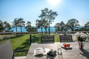 Ein Tisch mit einer Zeitschrift, Ferngläsern, Äpfeln und einem Glas Wasser, mit Blick auf eine Wiese und das Meer.