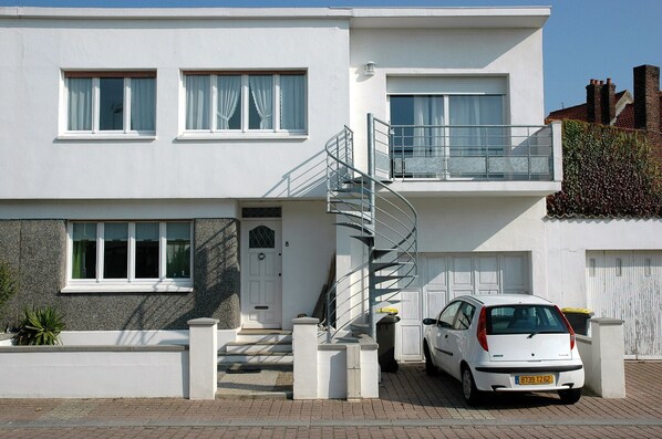 Une maison moderne à deux étages, avec une façade blanche, une entrée principale, un escalier et une voiture blanche garée devant.