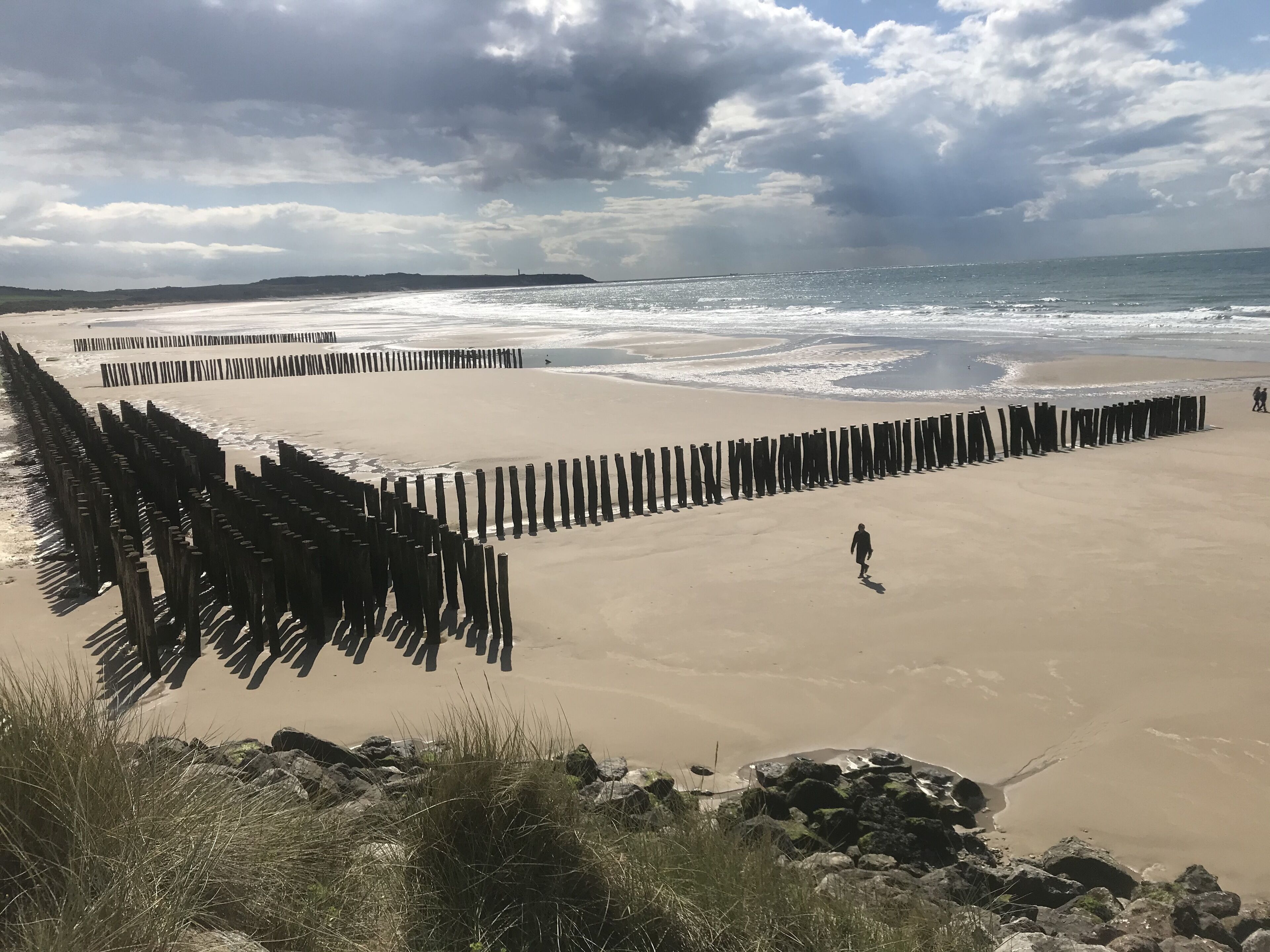 Een strand met een houten steiger en iemand die over het zand loopt.