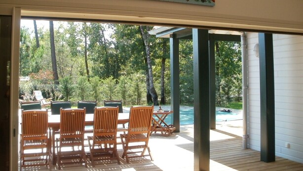 An open patio with a wooden table and chairs, a swimming pool, and a view of a forested area.