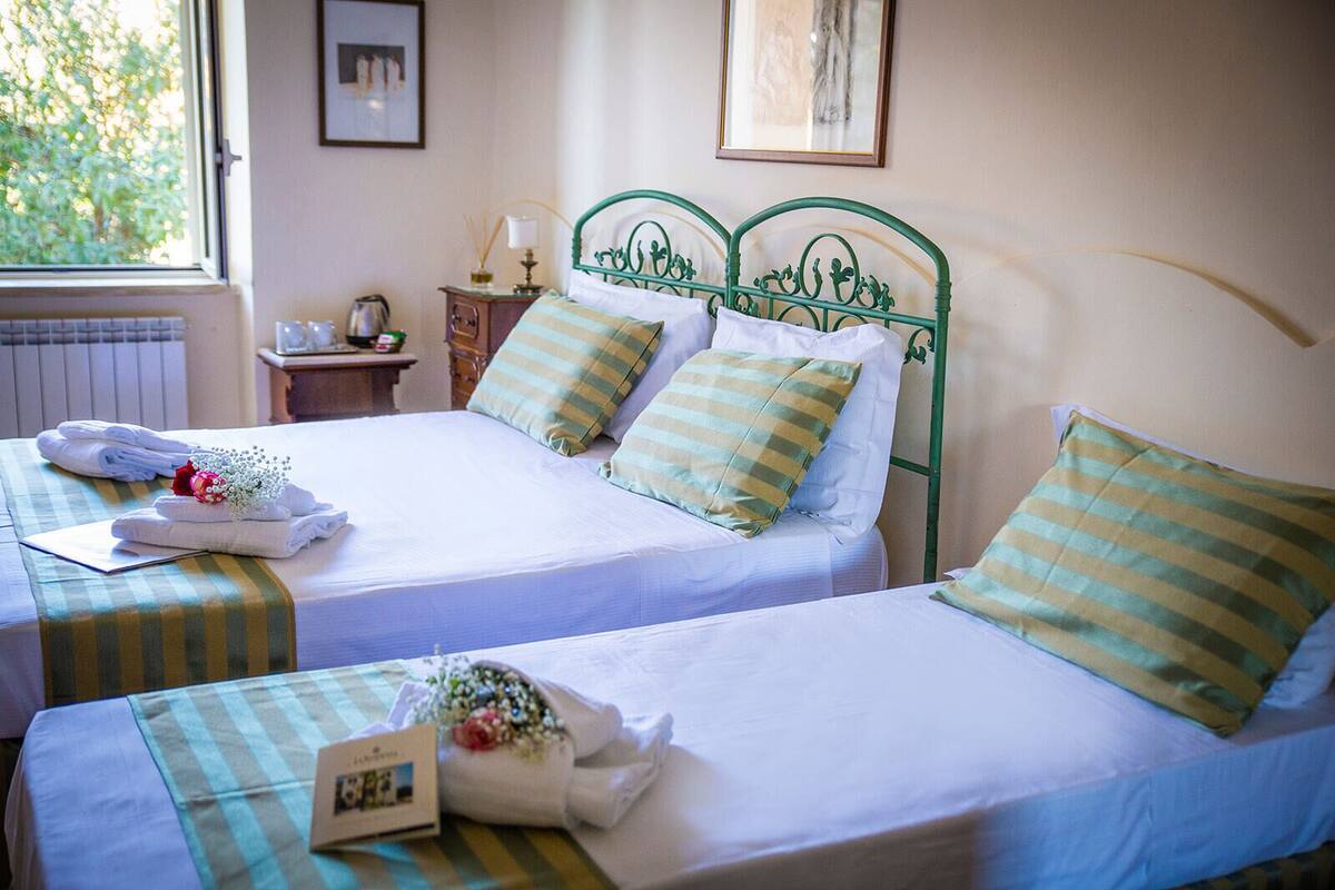 A hotel room with two beds, each with a green metal headboard, white linens, and striped pillows. There is a small bedside table with a lamp and a picture frame. A window provides a view of trees outside.