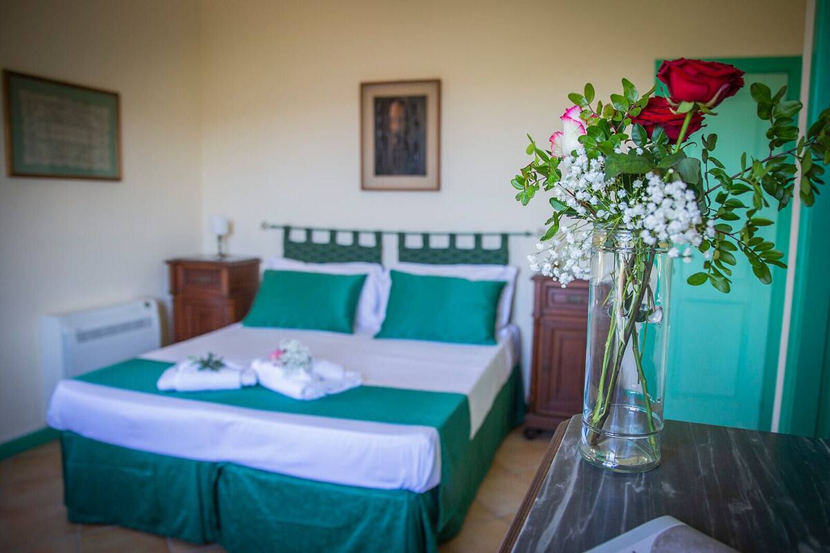 A neatly made bed with green bedding, a vase with flowers, and a red rose on a table.