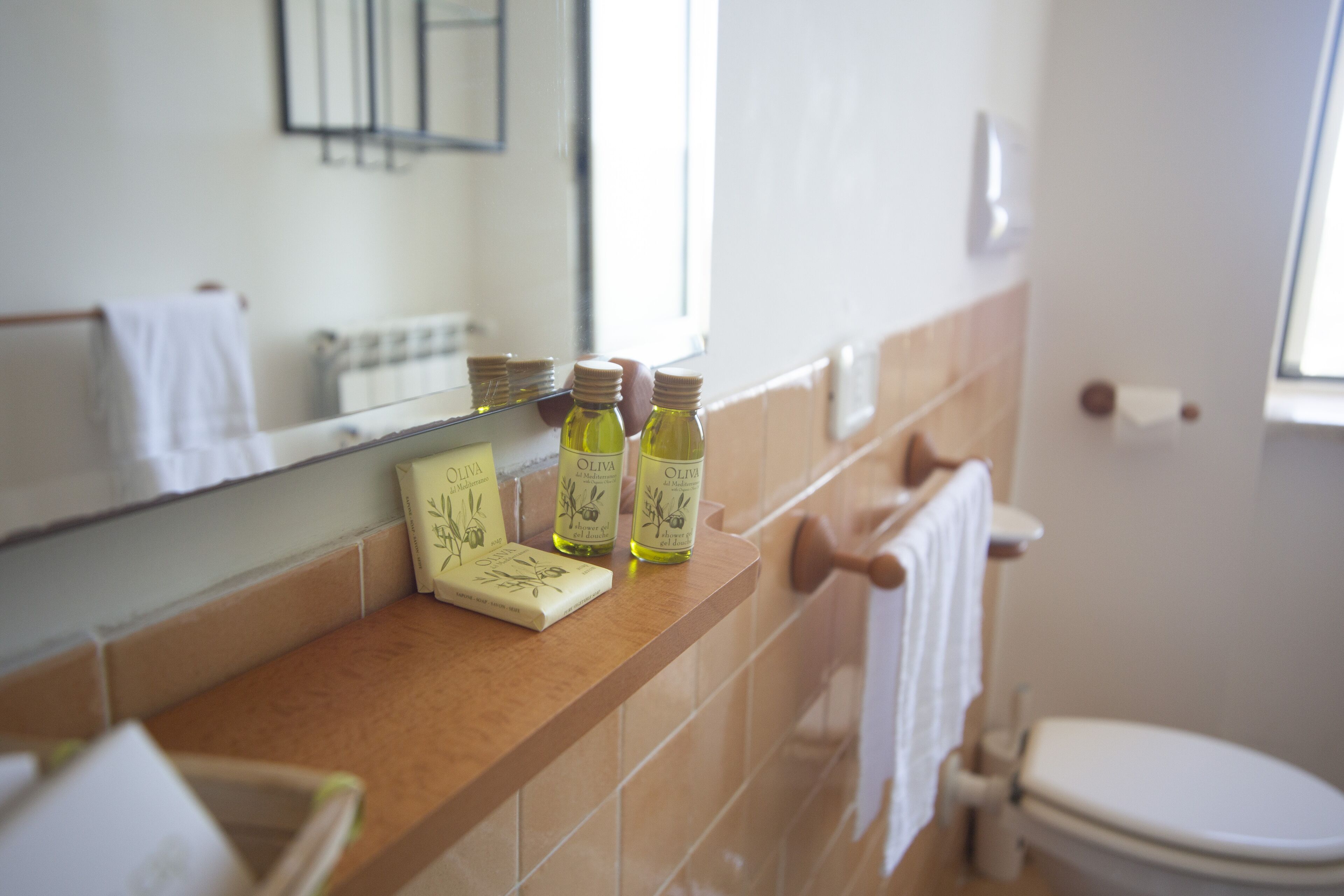 A bathroom with a beige countertop, a mirror, and a white toilet. There are three bottles of olive oil on the counter.