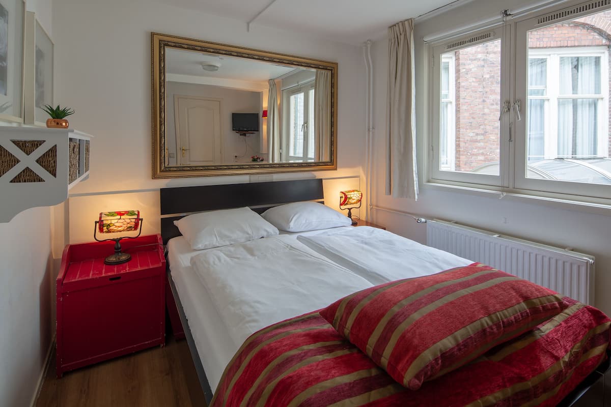 A neatly made bed with white linens and striped pillows, a red bedside table with a lamp, a large mirror above the bed, and a window with curtains.