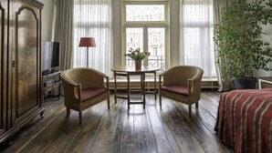 A room with a wooden floor, two wicker chairs with red cushions, a small round table with a vase of flowers, a television on a cabinet, and a large potted plant. - Amsterdam House Hotel (Amsterdam)