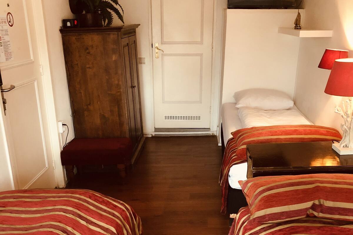 A hotel room with three single beds, wooden flooring, a white door, a cabinet, and a TV mounted on the wall.