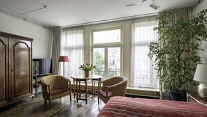 A hotel room with a bed, two wicker chairs, a small table with a vase, a television, and a large window with curtains. - Amsterdam House Hotel (Amsterdam)