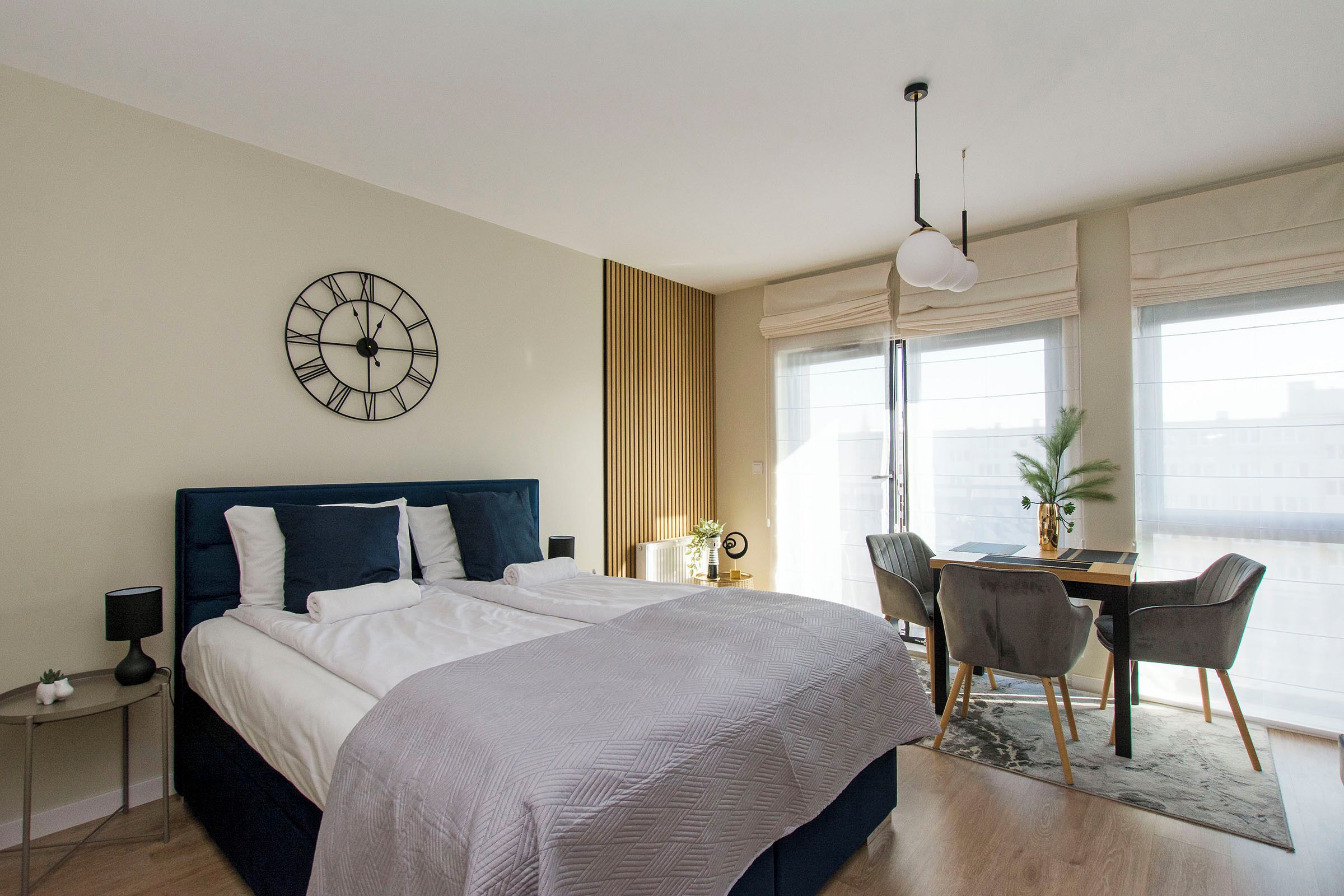 A modern bedroom with a large bed, a desk with two chairs, a wall clock, and a window with blinds.