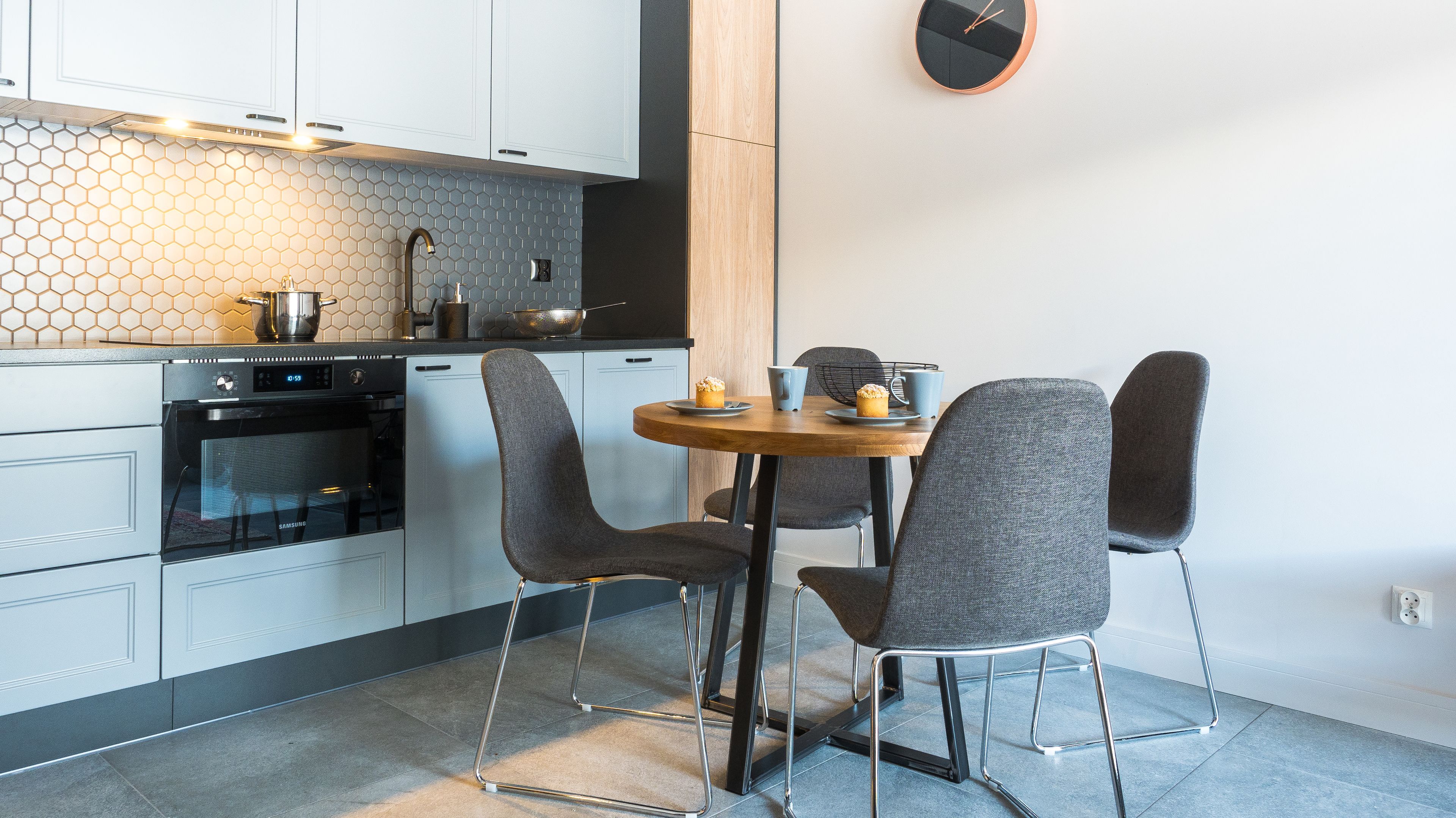 A modern kitchen with white cabinets, a black countertop, and a hexagonal backsplash. There is a round wooden table with four grey chairs.